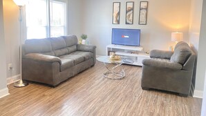A living room with a grey sofa, a coffee table, and a flat-screen TV on a white stand. There are three framed pictures on the wall. - Spacious Townhouse. 3B & 1. 5 baths. 10 Min to Airport. Free parking. (North Charleston)