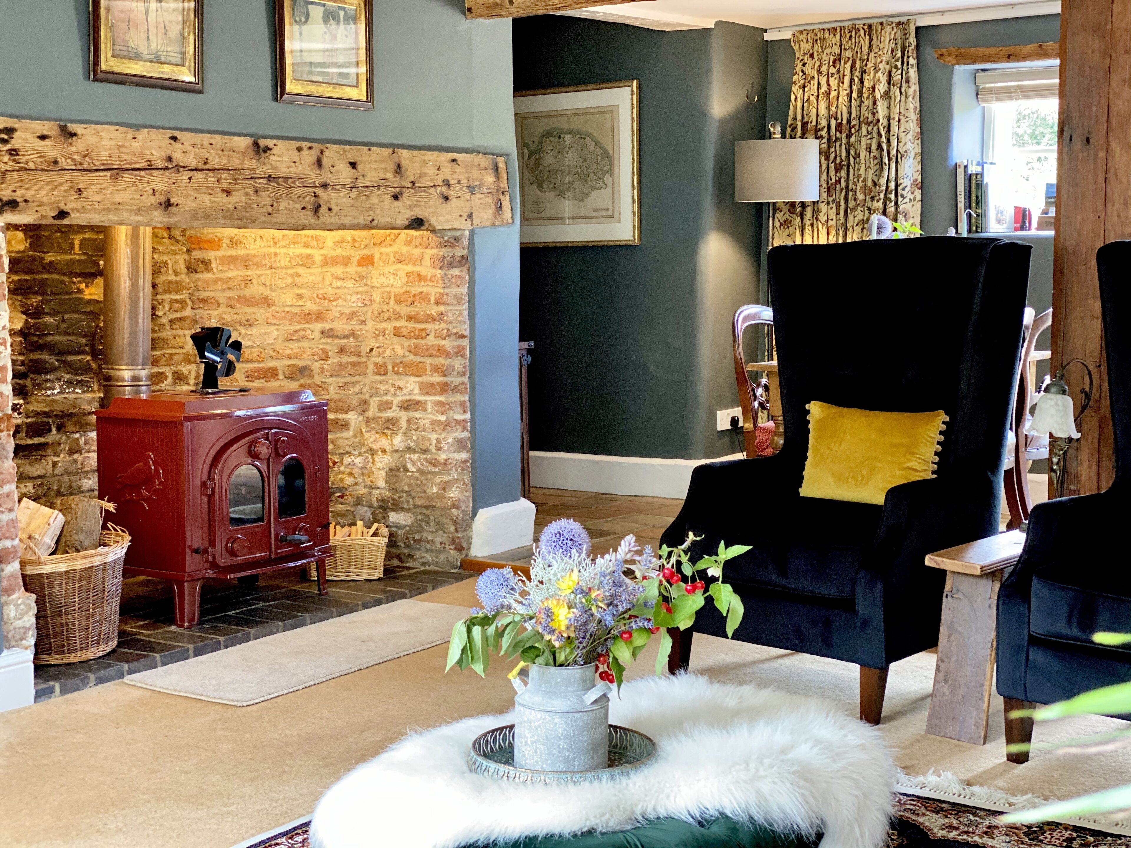 Una sala de estar acogedora con una chimenea de ladrillo, un sillón negro con un cojín amarillo y un centro de mesa floral sobre una mesa redonda.