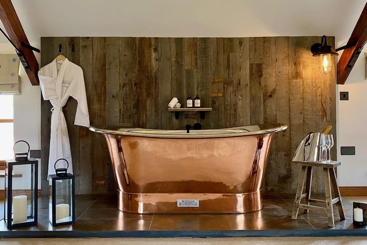 A spacious bathroom with a copper bathtub, wooden wall paneling, a robe hanging on a rack, and a shelf with toiletries.