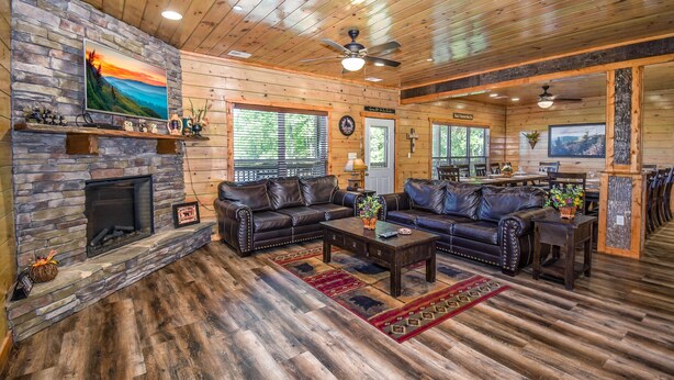 A spacious living room with a stone fireplace, leather sofas, and a dining area with a view of the outdoors. - SmokyStays 10 Bedroom Cabin (Pigeon Forge)
