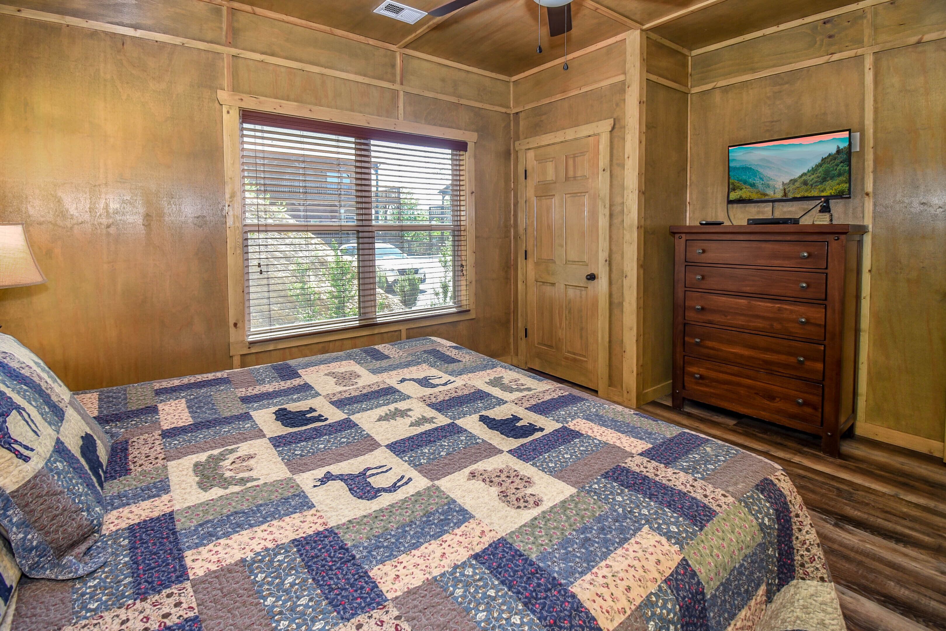 A bedroom with a wooden ceiling, a large window with blinds, a bed with a quilt, a dresser, and a television on a stand.
