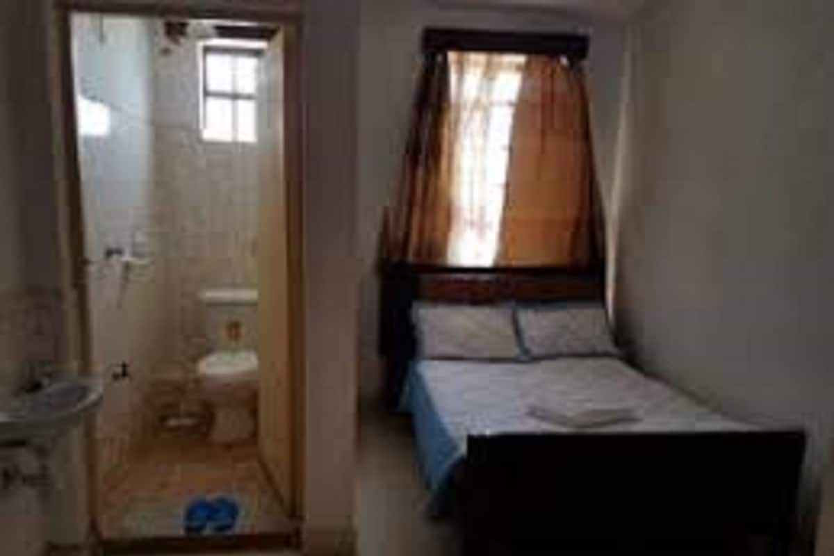 A small, narrow room with a single bed, a window with curtains, a sink, and a toilet.