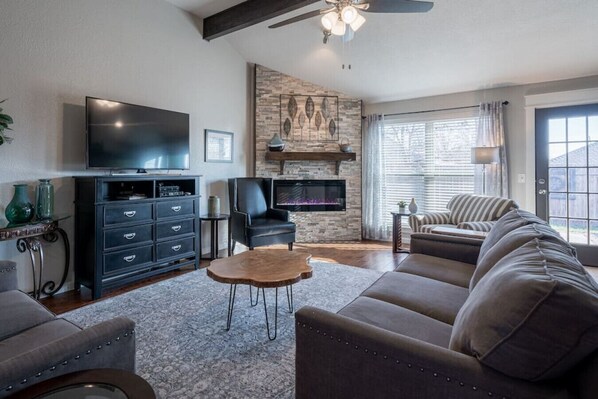 A modern living room with a flat-screen TV, leather sofas, a fireplace, and a large window with blinds.