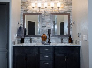 A bathroom with a stone backsplash, dual sinks, and dark cabinetry. - Fox Hunt · Modern, Updated Home · Pet-Friendly (Edmond)