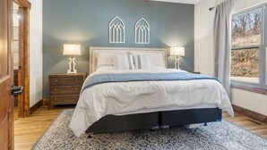 Une chambre à coucher comprenant un lit, deux tables de chevet, un ventilateur de plafond et une fenêtre avec des rideaux.