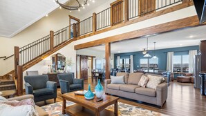 A spacious living area with a high ceiling, wooden beams, a staircase, and a view of the kitchen and dining area. - Lake View Escape - Sleeps 22, Game Room, Indoor Pool (Branson)