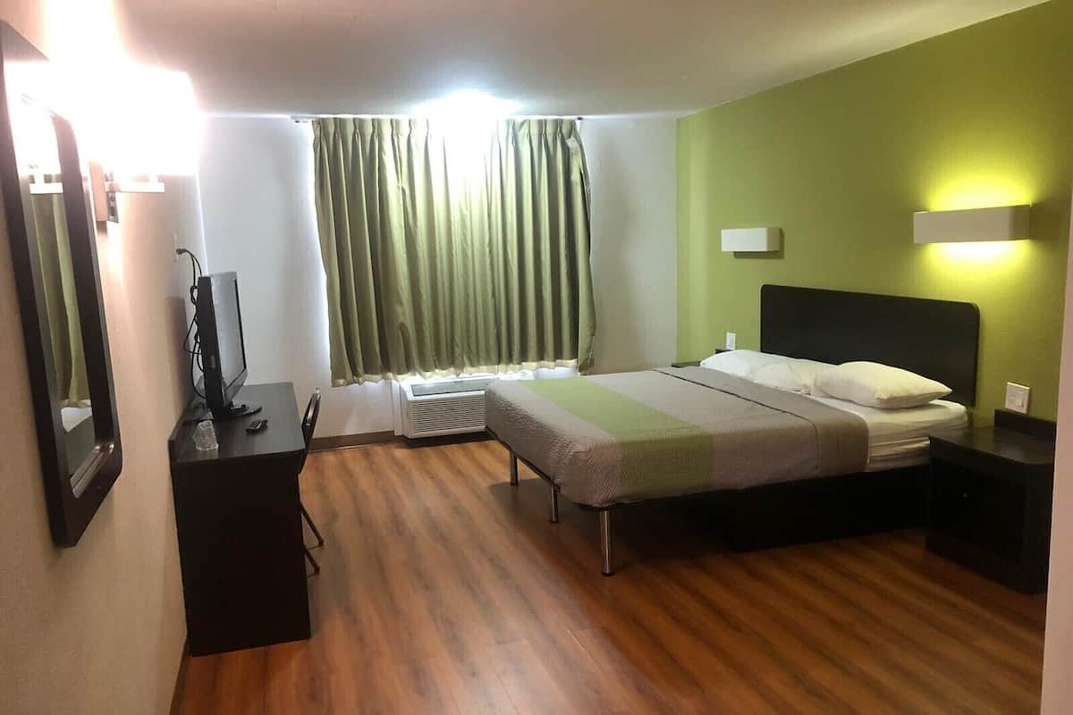 A hotel room with a bed, bedside table, a desk with a computer, a mirror, and a window with curtains.