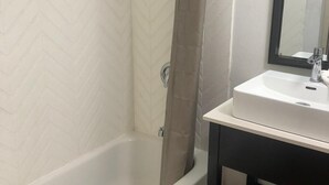 A bathroom with a white herringbone-patterned wall, a white bathtub, a glass shower enclosure, and a black vanity with a white sink. - Shadeland Inn Indianapolis (Indianapolis)