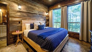 Een slaapkamer in een houten hutje, met een groot bed, een klein tafeltje en een raam met gordijnen.