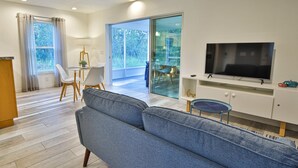 En moderne stue med en grå sofa, et fjernsyn på et hvidt skab, et spiseområde med bord og stole, og en skydedør til en altan.