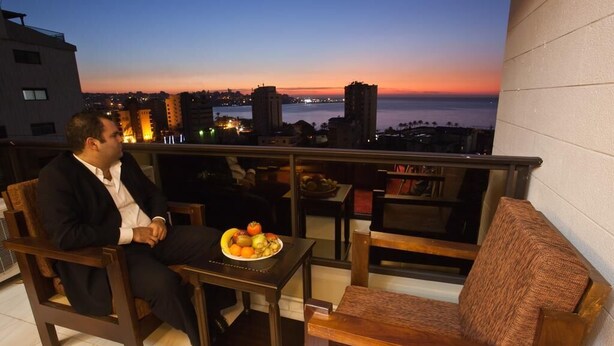 Ein Mann sitzt auf einem Balkon mit Obst auf dem Tisch und blickt auf einen Sonnenuntergang über der Skyline der Stadt.