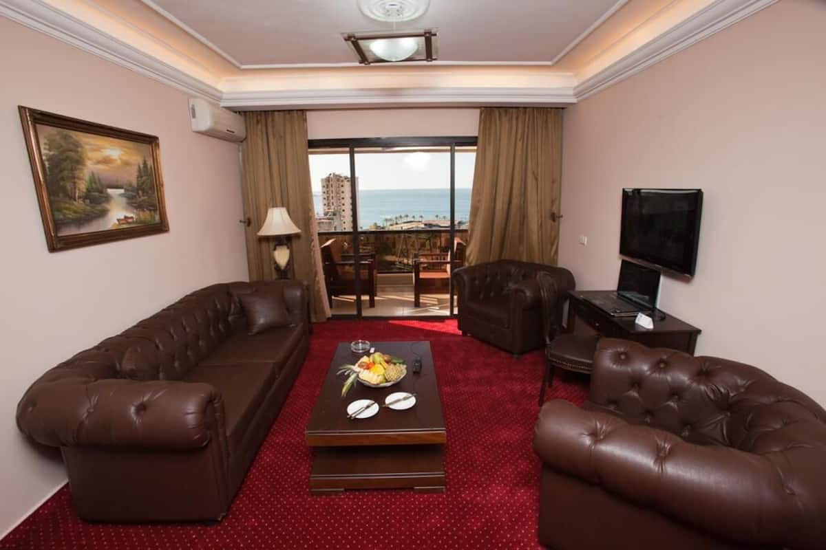 A hotel room with a red carpet, two leather sofas, a coffee table with fruit, a TV, and a painting of a landscape.