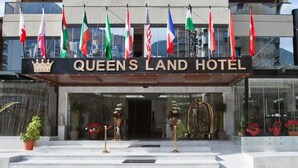 Eingang des Queens Land Hotels mit mehreren Flaggen, einem Kronenwappen und Topfpflanzen.