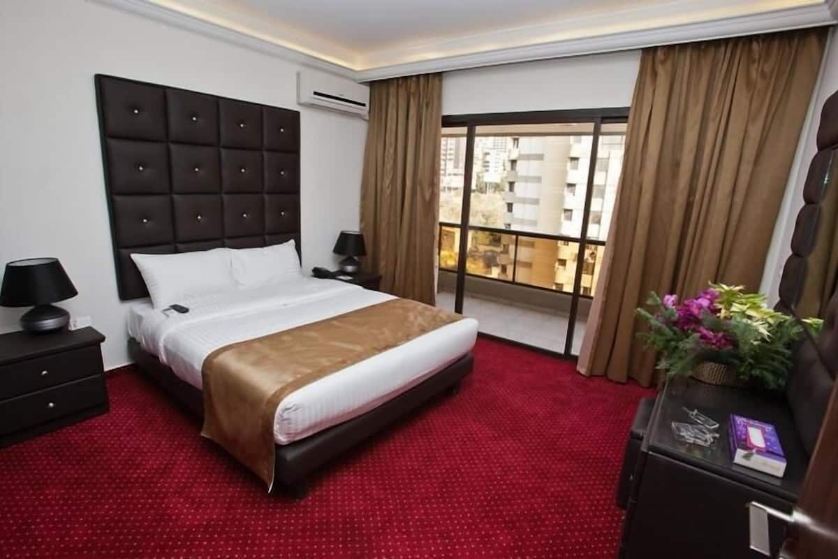 A hotel room with a large bed, a nightstand with a lamp, a dresser, and a view of the city through a large window.