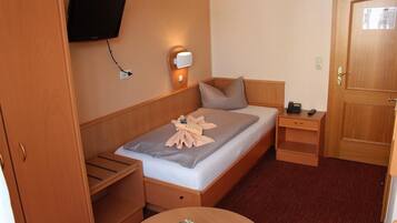 A compact hotel room with a single bed, a bedside table, a small table with a decorative item, a wall-mounted TV, and a door.