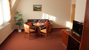 A cozy living room with a brown leather sofa, a wooden coffee table, and two armchairs. A potted plant is in the corner, and a painting hangs on the wall.