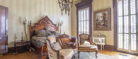 A bedroom with a large bed, two armchairs, a chandelier, and a painting on the wall.
