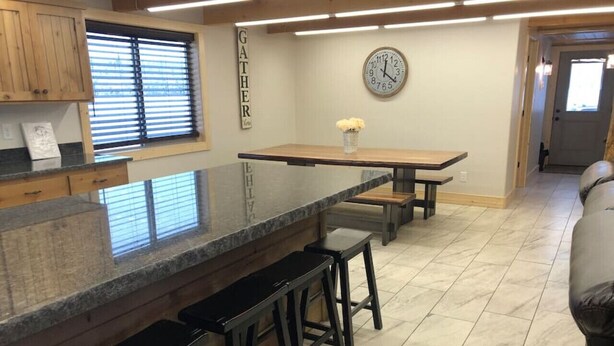 Une cuisine avec un îlot central, des tabourets de bar, une horloge et une fenêtre avec des stores.