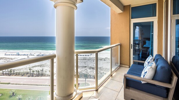Ein Balkon mit Blick auf Strand und Meer, eine Couch mit Kissen und eine Glastür zu einem Innenraum.