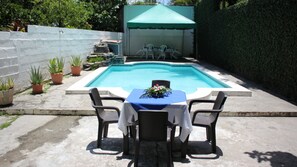 A pool with a table and chairs in the foreground, a covered area with chairs in the background, and a white wall on the left.