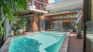 Un espace piscine extérieur moderne, doté d’une terrasse en bois, d’un patio vitré et d’une végétation luxuriante.