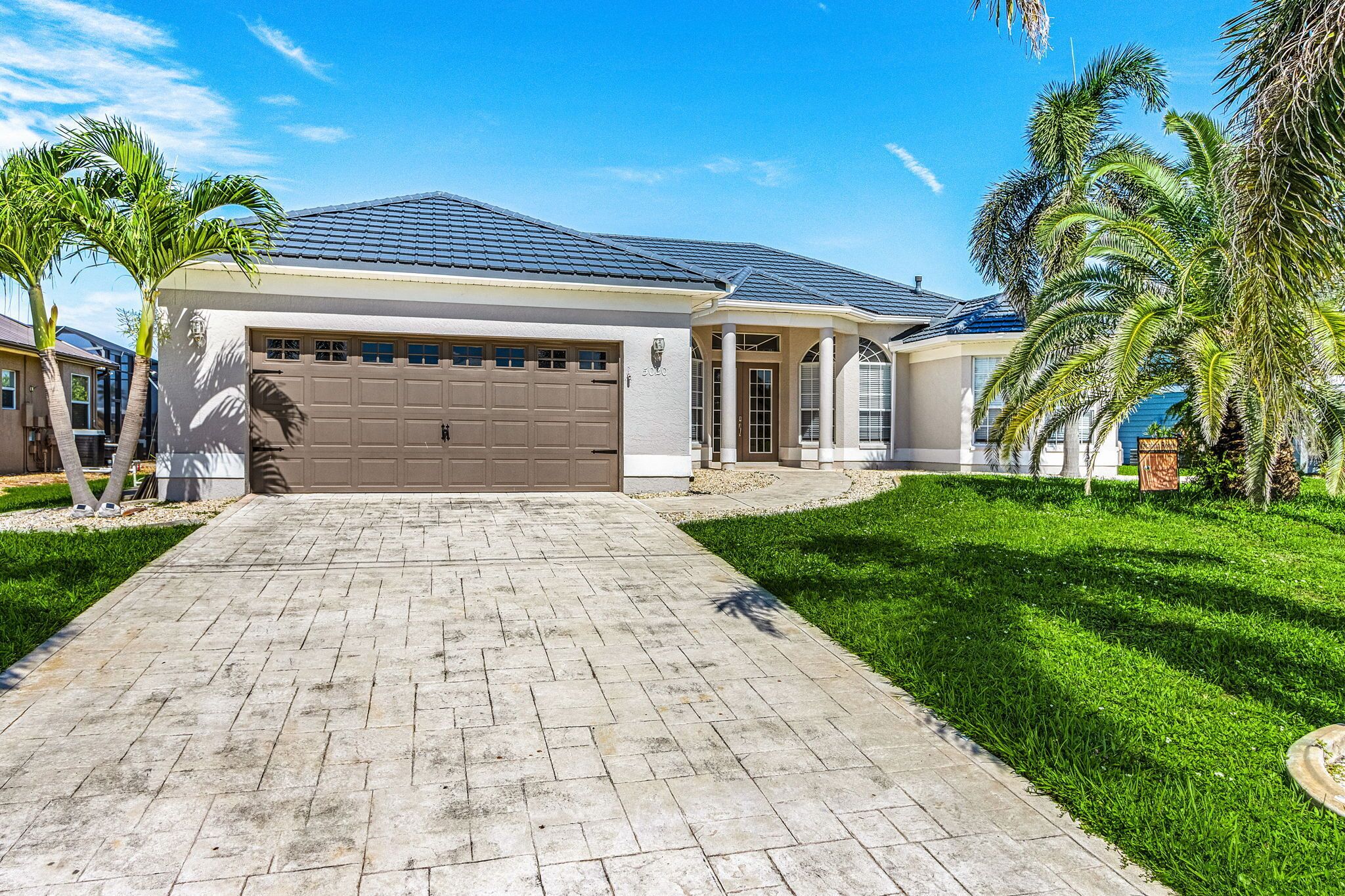 A single-story house with a garage, a paved driveway, and a well-maintained lawn with palm trees.