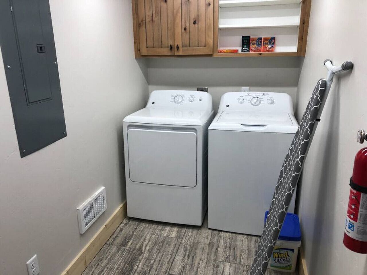 A laundry room with two white washing machines, a wooden cabinet above, a fire extinguisher, and a vent on the wall.