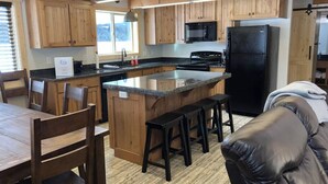 Una cocina moderna con armarios de madera, una isla central con taburetes, un refrigerador negro y un comedor con sillas de madera.