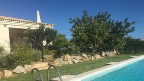 A swimming pool with a stone border, a metal ladder, and a building with a white facade and a tower in the background. - House - Holiday Home (Olhão)