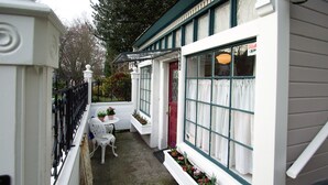 Un estrecho pasillo exterior con paredes blancas, una puerta roja y ventanas con cortinas blancas. Hay sillas exteriores blancas y macetas con flores.