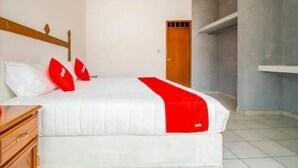 Un dormitorio con cama, cabecero de madera, paredes blancas, una puerta de madera y suelo de baldosas.