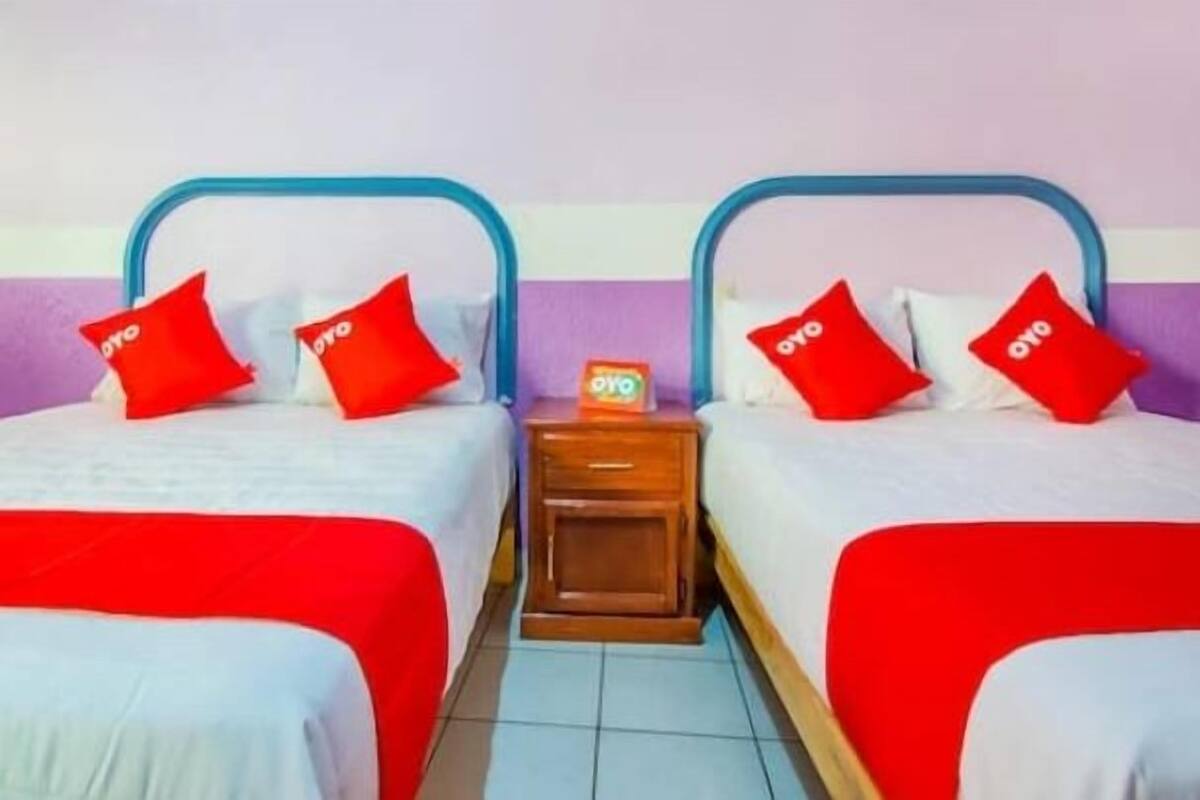 A hotel room with two beds, each with a blue metal headboard, red bedding, and matching red pillows. A wooden nightstand with a small table and a red object on it is between the beds.