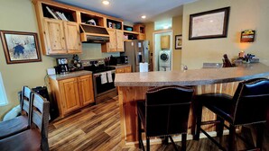 A kitchen with wooden cabinets, a stainless steel stove, a microwave, and a sink. There are two bar stools and a framed picture on the wall. - Glacier Corner: Enhance Your Adventure (East Glacier Park Village)