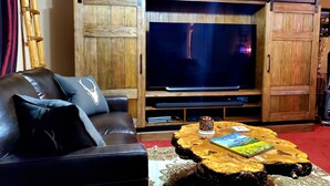 A living room with a black leather sofa, a wooden entertainment unit with a flat-screen TV, and a coffee table with a book. - Glacier Corner: Enhance Your Adventure (East Glacier Park Village)