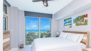 Une chambre dotée d’un grand lit, d’un ventilateur de plafond et offrant une vue sur l’océan grâce à de grandes fenêtres du sol au plafond.