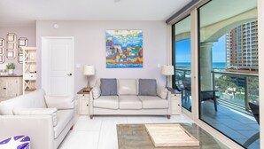 Un salon avec un canapé blanc, un divan beige, une table basse et un tableau accroché au mur. Il y a un balcon d’où l’on peut admirer un immeuble et l’océan.