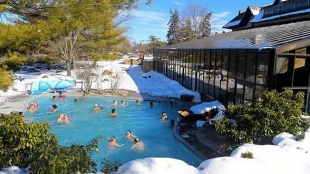 Una piscina rodeada de nieve, con gente nadando y un edificio al fondo.