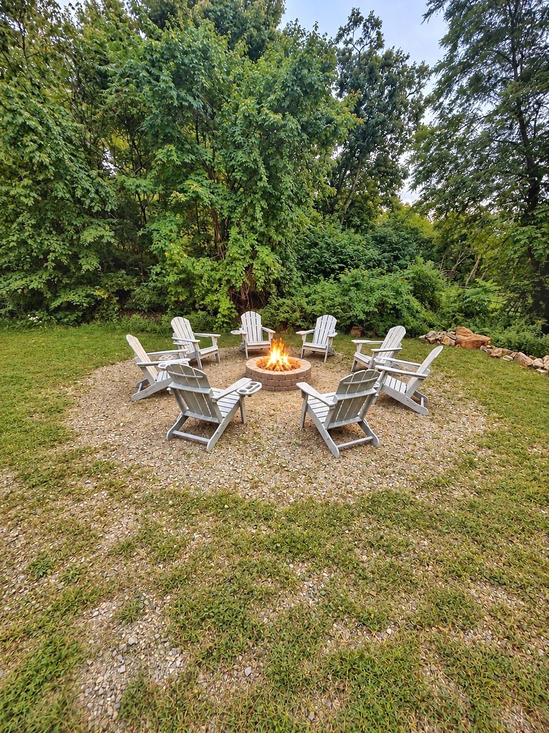 A fire pit surrounded by six Adirondack chairs in a grassy outdoor area with dense trees in the background.