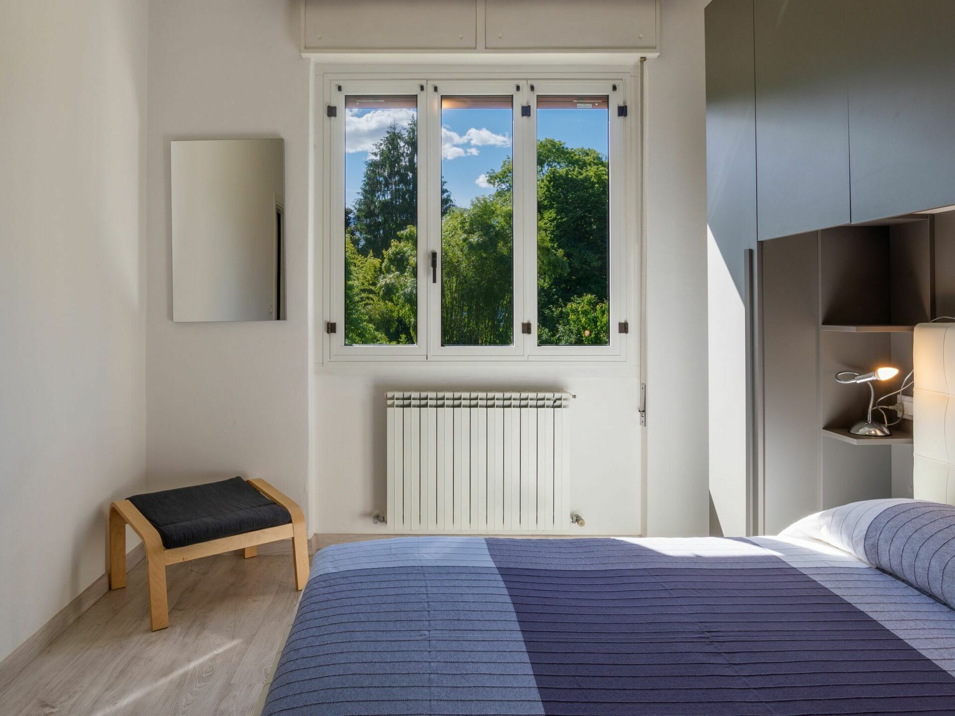 A bedroom with a large bed, a bench, a radiator, a window with a view of trees, and a desk with a lamp.