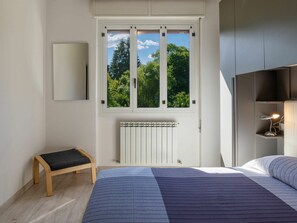 Ein Schlafzimmer mit einem großen Bett, einer Bank, einem Heizkörper, einem Fenster mit Blick auf Bäume und einem Schreibtisch mit Lampe.