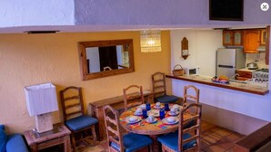 Uma área de jantar com uma mesa redonda posta para quatro pessoas, rodeada por cadeiras de madeira com almofadas azuis. Um pequeno criado-mudo de madeira com uma lâmpada está à esquerda. O espaço possui um piso de azulejos e uma cozinha ao fundo, com geladeira e fogão.