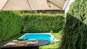 Un espacio junto a la piscina con una mesa lista para comer, una alberca y una sombrilla grande que da sombra.