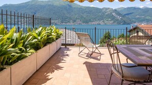 A terrace with a view of a lake and mountains, featuring a yellow striped awning, a chair, and a table.