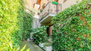 A narrow alleyway with a cobblestone path, surrounded by lush greenery and a building with a balcony.