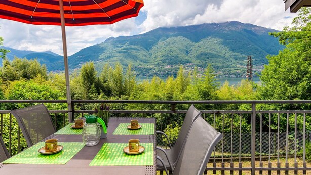 A balcony with a table set for two, a striped umbrella, and a view of mountains and a lake. - Beautiful private villa for 7 guests with WIFI, TV, terrace and pets allowed (Vestreno)