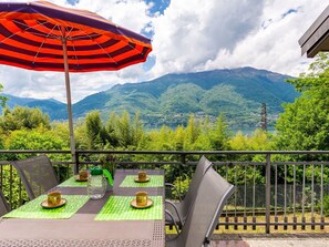 En balkon med et bord dækket til to, en stribet parasol og udsigt til bjerge og en sø.