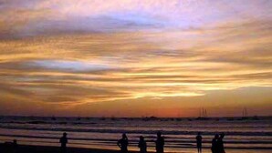 Une scène de plage avec des silhouettes de personnes et des voiliers, le tout baigné par la lumière d’un coucher de soleil.
