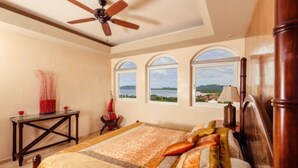 Une chambre à coucher comprenant un lit, un ventilateur de plafond, une table de chevet et trois fenêtres donnant sur la mer.