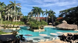Un espace piscine aménagé dans un complexe hôtelier, avec des palmiers, des chaises longues et des parasols.