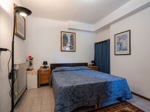 Un dormitorio con cama, dos mesitas de noche, un radiador, lámpara, un cuadro enmarcado y una cortina.